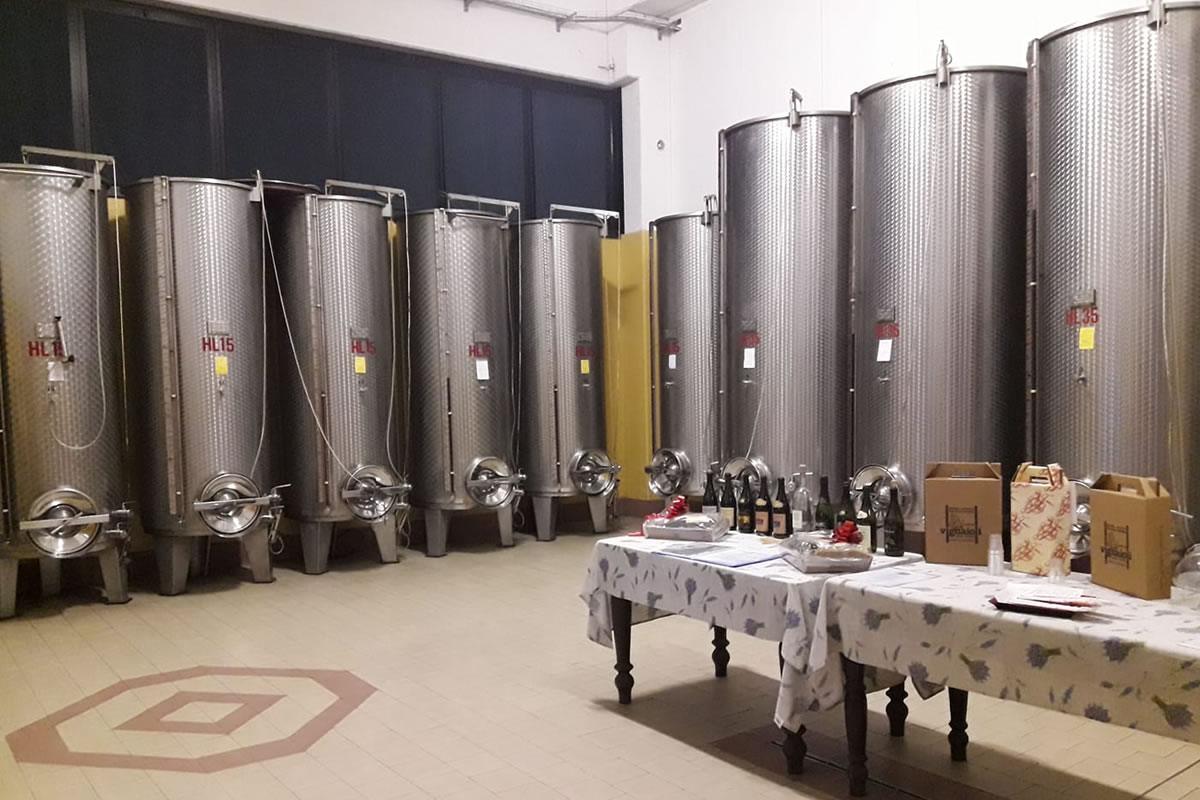 Cantina vinicola con serbatoi in acciaio inox. Tavoli con bottiglie e confezioni di vino. Pavimento con decorazione geometrica.