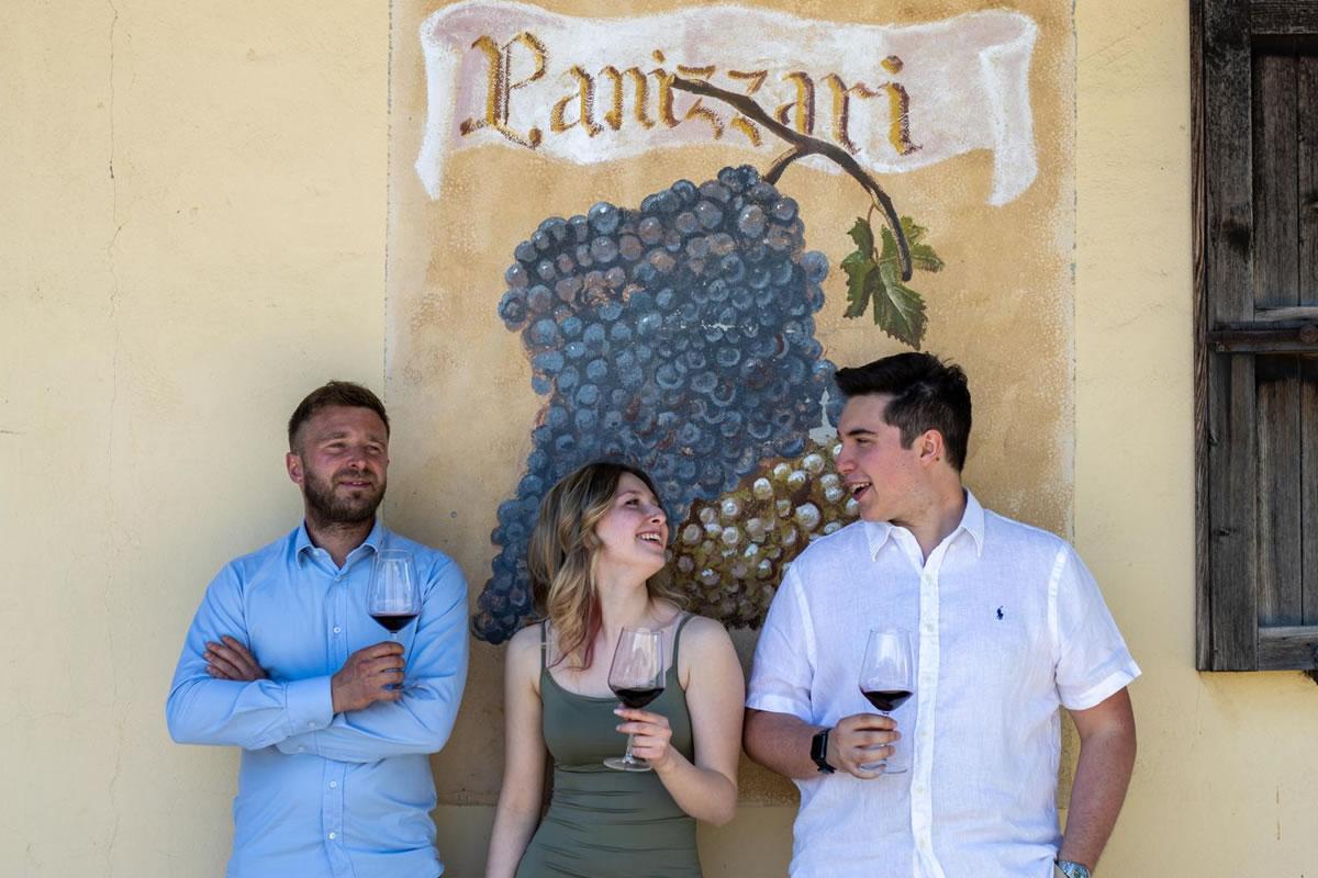 Tre persone sorridono con bicchieri di vino rosso davanti a un murale con uva e la scritta Panizzari. Azienda agricola.