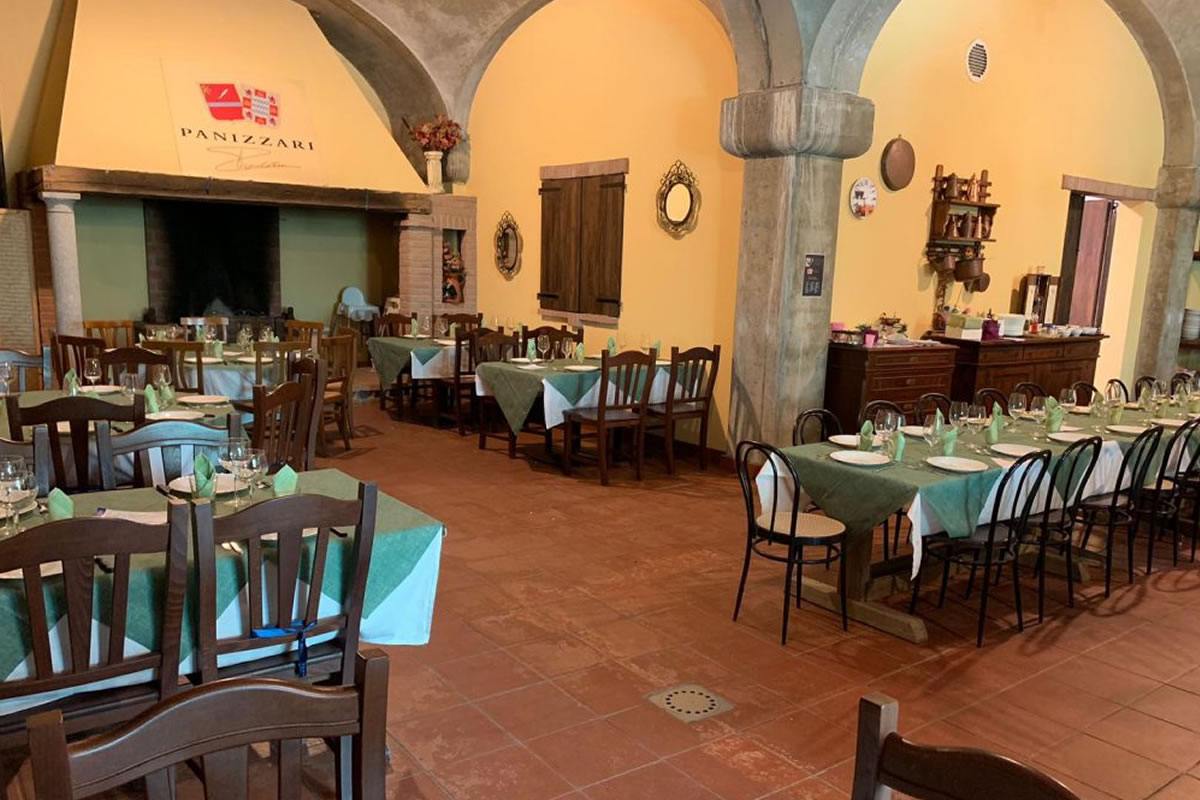 Interno ristorante Panizzari con tavoli apparecchiati, sedie in legno, camino, colonne e soffitto a volta. Atmosfera rustica e accogliente.