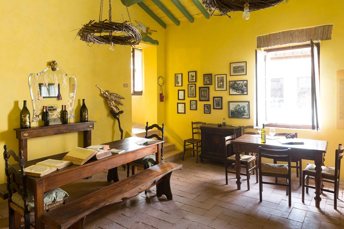 Interno rustico con pareti gialle. Tavoli e sedie in legno, bottiglie di vino e decorazioni. Soffitto con travi a vista e lampade.