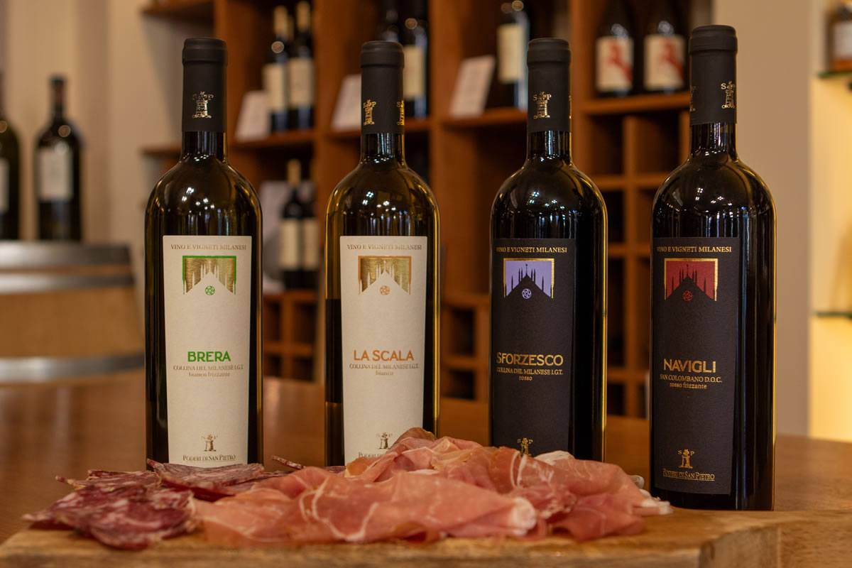 Quattro bottiglie di vino rosso Poderi San Pietro con salumi su tagliere in legno. Etichette: Brera, La Scala, Sforzesco, Navigli.