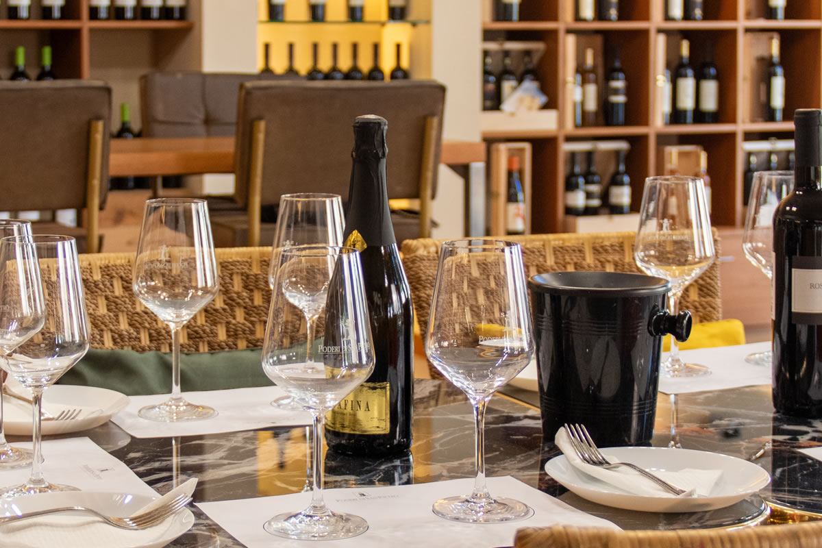 Tavola imbandita per degustazione vini: calici, bottiglie, secchiello ghiaccio. Sfondo con scaffali di bottiglie e sedie in vimini.