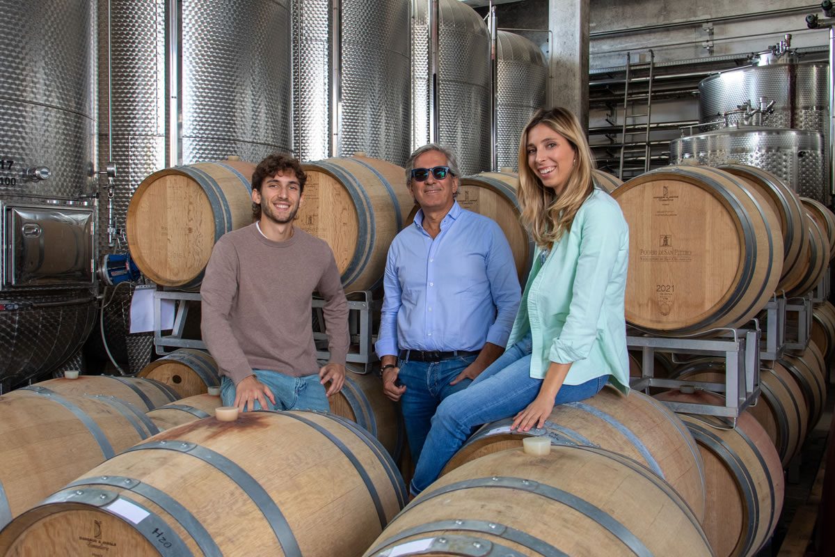 Tre persone in una cantina vinicola con botti di legno e serbatoi in acciaio. Una donna seduta su una botte.